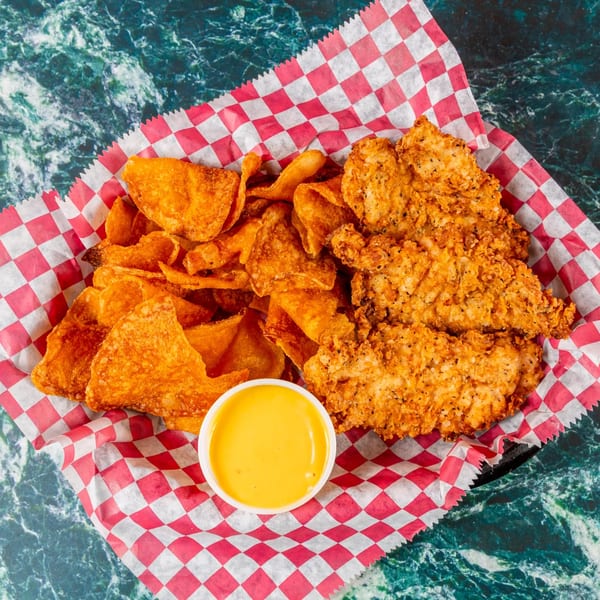 Fried chicken tenders, golden chips, and a small cup of dipping sauce in a red and white checkered basket.