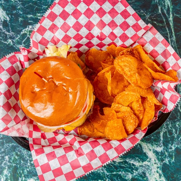 A burger with cheese, onions, and pickles, served with potato chips in a red and white checkered basket.